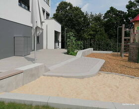 Spielplatz mit Sandkasten, Klettergerüst, Kies, Terrasse und einer grauen Box vor einem Gebäude, umgeben von Bäumen.