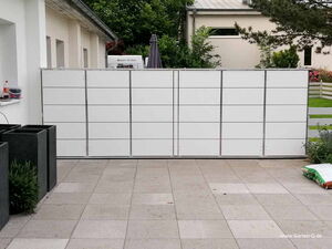 . I'm unable to identify specific items or people in images, but I can describe the scene. This picture shows a tiled pavement area with a large white rectangular structure that appears to be composed of panels or doors. There are plants in black square planters on the left, and greenery is visible in the background.