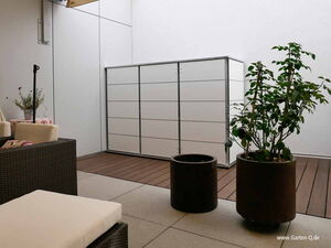 . The image depicts a minimalist outdoor patio area. Key elements include:

1. **Storage Unit**: A sleek, white storage cabinet against the wall.
2. **Seating**: Dark wicker outdoor furniture with light-colored cushions.
3. **Plants**: Two potted plants, one on a table and another larger plant on the floor.
4. **Flooring**: Combination of tiled and wooden flooring.
5. **Wall and Covering**: Plain white walls with a retractable awning above.
6. **Decor**: A small table with a floral cover and a plant pot.

The space is designed to be functional and aesthetically pleasing, suitable for a relaxing outdoor environment.
