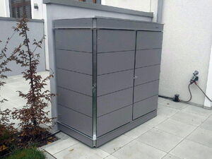. Ein grauer Metallschrank steht auf einer Terrasse neben einem kleinen Baum, mit einem Wasserschlauch in der Nähe.