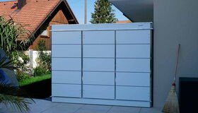 . The image shows a white metal storage unit or cabinet located outdoors. It's placed against a wall on a patio or tiled surface. There's a broom leaning against the wall nearby, and part of a chair or similar object is visible. In the background, there is a wooden house with a red roof and some greenery.