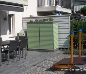 . The image shows a patio area with several elements:

1. A dining table with chairs made of a dark material.
2. A green garden storage unit with a planter on top, containing some plants.
3. The exterior of a house or building with windows and a railing above.
4. A small wooden structure with strings and blue elements, possibly part of a game or garden decor, on the right.
5. Paved and gravel ground surfaces.
6. Nearby greenery and shrubs.

Overall, it appears to be an outdoor living area with functional and decorative elements.