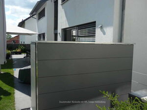 . The image shows a modern residential outdoor area. There is a patio with tables and chairs, a large umbrella, and a sleek structure with horizontal panels, possibly a storage unit or decorative wall. The area is surrounded by a building, some greenery, and paving stones. The house has a white exterior with some windows.