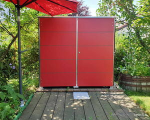 Rotes Gartenhäuschen auf Holzterrasse, umgeben von Büschen, mit rotem Sonnenschirm links.