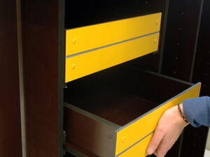 . Ein grauer Schrank mit zwei gelben Schubladen. Eine Hand öffnet die untere.