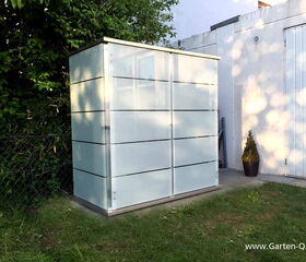 . The image shows a modern, rectangular outdoor storage shed located in a garden area. It appears to be made of metal or a similar material, with horizontal lines and a sleek design. The shed is placed near a fence, surrounded by greenery, and there's a concrete pathway beside it. A potted plant is visible near a wall to the right.