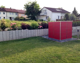 . The image shows a suburban scene with a small yard area. In the foreground, there's a well-maintained lawn. To the right, there is a red, cube-like structure, possibly a small shed or enclosure. Behind it is a row of concrete blocks or a low stone wall that separates the lawn from a garden area. The garden has shrubs and blooming roses. In the background, there are residential houses with sloped roofs, some with skylights and balconies. Trees and more shrubs are visible, adding greenery to the scene.