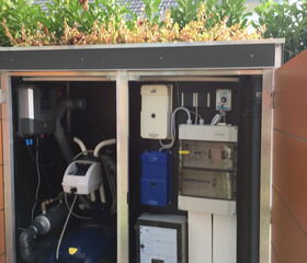 The image shows the interior of a technical cabinet for a swimming pool system. Inside, there is pool equipment such as pumps, filters, control units, and possibly a robotic pool cleaner. The cabinet is located outdoors and is topped with some greenery, likely as part of a green roof system for aesthetic purposes.