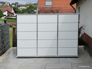 . Ein moderner, weißer Gartenschrank auf einer Terrasse, mit einem Hintergrund aus Häusern und Bäumen.