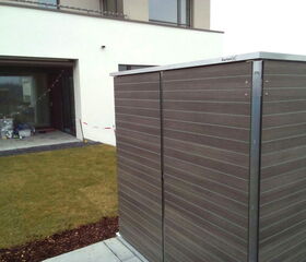 . Ein Außenbereich mit Rasen, Terrasse und Gartenbox neben einem modernen Gebäude.
