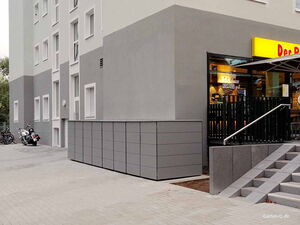 . Ein graues Gebäude mit modernen Mülltonnenboxen daneben und ein Geschäft mit gelbem Schild rechts.