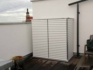 . Ein Balkon mit weißem Schrank, Pflanzentöpfen und einer Kirchturmspitze im Hintergrund.