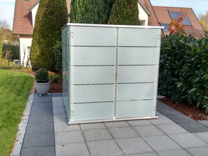 . The image shows a frosted glass enclosure placed on a tiled patio area. There's a potted plant on the left, and the surroundings include manicured shrubs and trees, a lawn, and some residential buildings in the background.