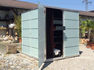 . This image shows an outdoor storage structure with its door open. The structure has a modern design with glass panels and appears to have shelves inside. There are various garden elements around it, such as potted plants and decorative objects.