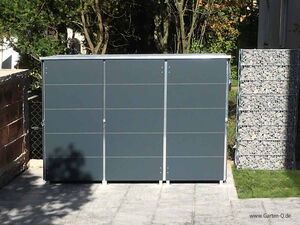 . The image shows a modern storage unit or garden shed with a sleek design, situated outdoors. It has a dark gray color with a metal frame, and it's positioned on a paved surface. To the right, there is a structure filled with stones, likely for decorative or structural purposes, and trees are visible in the background.