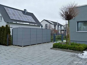 . Ein graues Gartenhaus vor einem modernen Haus mit Solarpanelen auf dem Dach, Pflastersteine und etwas Schnee.