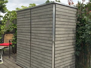 . Ein grauer Gartenschrank aus Holz auf einer Terrasse, umgeben von Pflanzen.