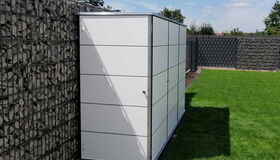 . Ein moderner, weißer Gartenschrank steht an einer Gabionenwand auf einer Rasenfläche.