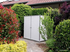 . The image shows a small, modern garden shed surrounded by various shrubs and greenery. The shed has a simple, sleek design with horizontal paneling and double doors. It's set in a landscaped garden area with different types of plants and bushes of varying colors and sizes. The background includes a house with a tiled roof.