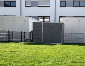 The image shows a modern-looking outdoor storage unit or shed, situated in a fenced area with a grassy lawn. Behind it, there is a residential building with multiple windows. The shed appears to be made of grey panels.
