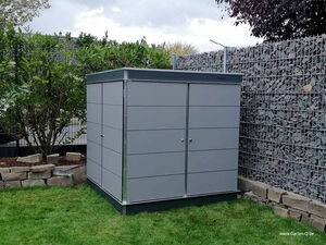 . Ein moderner, grauer Gartenschrank steht auf einer Wiese, umgeben von Pflanzen und Steinfassungen.