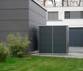 . This image features a modern, rectangular storage unit situated in a yard. It is adjacent to a contemporary-style building with clean lines and dark panels. There is some greenery, including grass and small bushes, in the foreground. The design is minimalist and sleek.