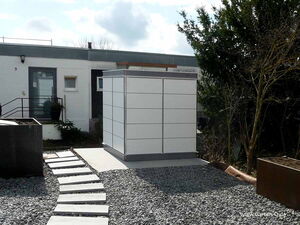. The image shows a modern outdoor storage unit or garden shed. It appears to be situated in a landscaped garden area, with a gravel path leading up to it. The unit is white and has a sleek, minimalist design. The surrounding area includes some potted plants and trees.