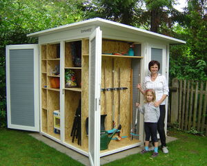 The image depicts a garden shed with open doors. Inside, there are various gardening tools, a basketball, a small play kitchen, and other items stored on shelves. Beside the shed, there are two people, an adult and a child, standing and smiling. The setting is in a garden with greenery around.