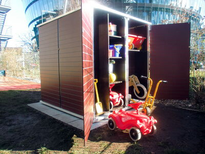 This image shows a storage shed with its doors open. Inside, there are various children's toys including:

1. A red toy car with "Bobby-Car" written on it.
2. A yellow toy wheelbarrow.
3. A balance bike.
4. A bucket and spade.
5. Several other toys and play equipment on shelves.

The shed is located outdoors, with grass and some modern buildings visible in the background.
