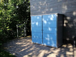 . Ein blauer Gartenschrank steht auf einer gepflasterten Fläche neben einem Gebäude, umgeben von Bäumen.