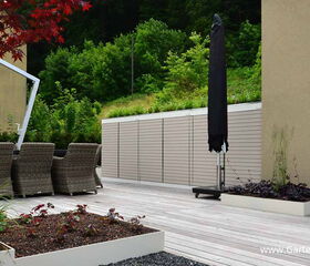 . This image shows a modern outdoor patio setting. There are several wicker chairs arranged around a table under an umbrella. The area is surrounded by lush greenery and a building with green shutters. There is also an umbrella stand, planter boxes with various plants, and a wooden deck. The environment suggests a garden or terrace setup designed for relaxation and outdoor dining.