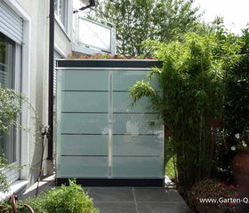 . The image shows an outdoor scene featuring a glass structure positioned against a house with a garden surrounding it. The structure has a modern design, likely used for storage or as a small shed. It is nestled among greenery, including bamboo and other plants in pots. The area appears well-maintained and landscaped.