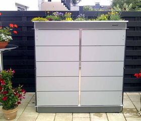 . The image shows an outdoor setting with a modern, sleek storage unit against a dark fence. The unit has a planter on top, filled with various colorful flowers. There are additional potted plants placed on either side of the storage unit, adding to the decorative greenery of the space. A small round table holds another potted plant on the left. Overall, the scene appears to be a landscaped garden or patio area.