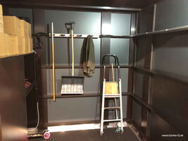 The image shows the interior of a storage shed or small workshop. Here's a description of the items present:

1. Cardboard boxes stacked on shelves.
2. A snow shovel and a broom hanging on the wall.
3. A jacket or garment hanging on a hook.
4. A small ladder or step stool.
5. Other tools like a hose or extension cord.
6. Metal shelves lining the walls.

The space appears organized and is likely used for garden or storage purposes.
