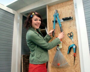 The image depicts a person organizing tools in a storage area. The tools visible include:

- Large pruning shears
- Hand saw
- Rake
- Hammer
- Scissors
- Gardening gloves
- Tape measure
- Small brush or tool with a blue handle

This person appears to be arranging or displaying these tools on the door of a storage cabinet.