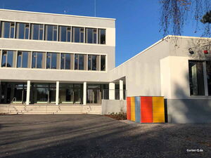 . Modernes Gebäude mit großer Glasfassade und buntem Container daneben auf einem sonnigen Freigelände.