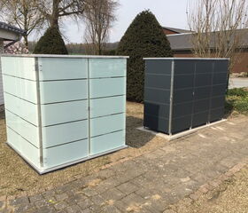 . Zwei moderne Mülltonnenboxen im Garten, eine aus weißem Glas, die andere in dunklem Grau.