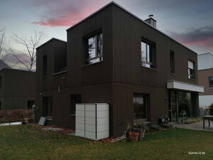. Moderne zweistöckige Häuser mit dunkler Fassade, Garten, Pflanzen und einem Schuppen im Vordergrund bei Sonnenuntergang.