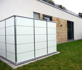 . The image shows a modern white utility enclosure or storage cabinet located outside a building. It has a sleek, panel design with a metal frame and is placed on a concrete slab. Nearby is a building with a mix of plaster and stone facade, and there are sliding glass doors visible. There’s also a lawn in the foreground.
