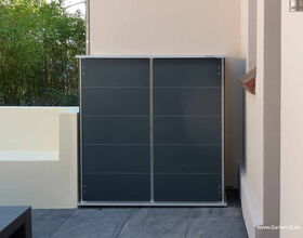 . The image shows a sleek, modern outdoor storage cabinet against a wall in a courtyard or garden area. It has a simple, flat design with smooth, dark gray panels. There is a patch of greenery in the background, suggesting the setting is part of a garden or patio.