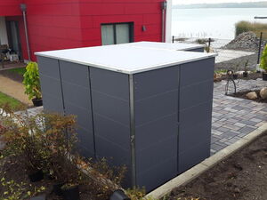 . The image shows an outdoor scene with a modern building featuring a red wall and black and gray elements. In the foreground, there's a cubic structure with a gray exterior, possibly a garden storage unit or shed. The area around it includes a paved walkway, plants, and landscaping work in progress. Nearby, there's a body of water and a person working on the landscaping.