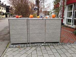 . The image shows a structure with a wooden panel exterior, possibly a bin or storage unit, placed along a sidewalk. On top of the structure, there are pumpkins and gourds arranged as decoration. The background features a street with a few buildings, trees, and a pathway. It appears to be a calm, outdoor urban setting.