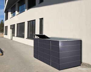 The image shows a large, modern, outdoor storage unit with a sleek design. It is placed against a building with multiple windows and blinds. The unit appears to be made of metal, with a dark color and a hinged lid on top that is partially open. The setting is an outdoor paved area next to the building.