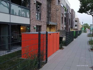 . The image shows a sidewalk next to a modern residential building with brick and glass exteriors. Along the path, there's a metal fence and a bright orange structure, possibly a utility box or storage unit. Trees and some greenery are visible, and the setting appears to be urban.