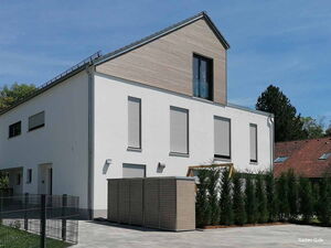 . Moderner weißer Bau mit Satteldach, großen Fenstern und Jalousien, umgeben von Bäumen und Hecken.