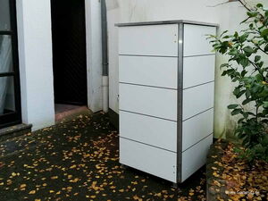 . Ein moderner grauer Außenlagerschrank steht auf einer Terrasse neben einer Wand und Sträuchern, umgeben von Herbstlaub.