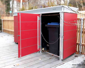. The image shows a red outdoor storage unit or shed with an open door, revealing a trash bin with a blue lid inside. The unit has a metal frame and a plant holder on top. It’s located on a wooden deck with a wooden fence in the background. There are some fallen leaves around.
