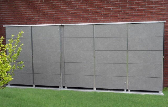 The image shows a set of gray storage cabinets or lockers placed outdoors. They are positioned against a brick wall, and there's some grass in the foreground along with a small leafy bush on the left side. The storage units appear to be made of metal or a similar material.