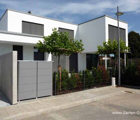 . The image shows a modern, minimalist two-story house with flat roofs and large windows covered with horizontal slats. In front of the house, there are neatly pruned trees, a gray wall, and a metal fence. There's also a streetlamp and a paved sidewalk bordering the property. The sky is clear and blue, suggesting a sunny day.
