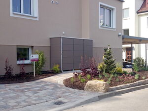 . The image shows a landscaped area in front of a residential building. Key elements include:

1. **Building Exterior**: A beige or light-colored building with several windows.
2. **Landscaping**: Various plants and shrubs neatly arranged in garden beds.
3. **Pathway**: A paved walkway made of stone or tiles leading to the entrance.
4. **Signage**: A signpost is present, though the text is not clear.
5. **Carport**: A covered parking area on the right side, partially visible.
6. **Rocks**: A few large decorative stones are part of the landscape design.

The overall scene suggests a well-maintained and aesthetically pleasing outdoor space.